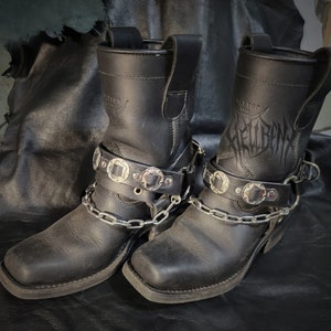 May include: A pair of black leather motorcycle boots with silver buckles and chains. The boots have the word "Hellbent" embossed on the side.