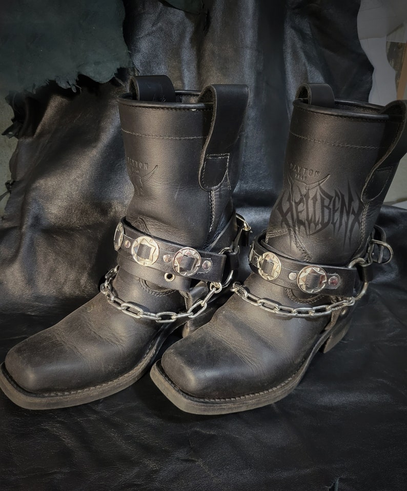 May include: A pair of black leather motorcycle boots with silver buckles and chains. The boots have the word "Hellbent" embossed on the side.