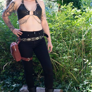 May include: A woman wearing a black leather halter top with a silver chain and ring detail. She is also wearing black jeans, a black belt with silver accents, a brown leather fanny pack, and black boots with silver chain and buckle accents.