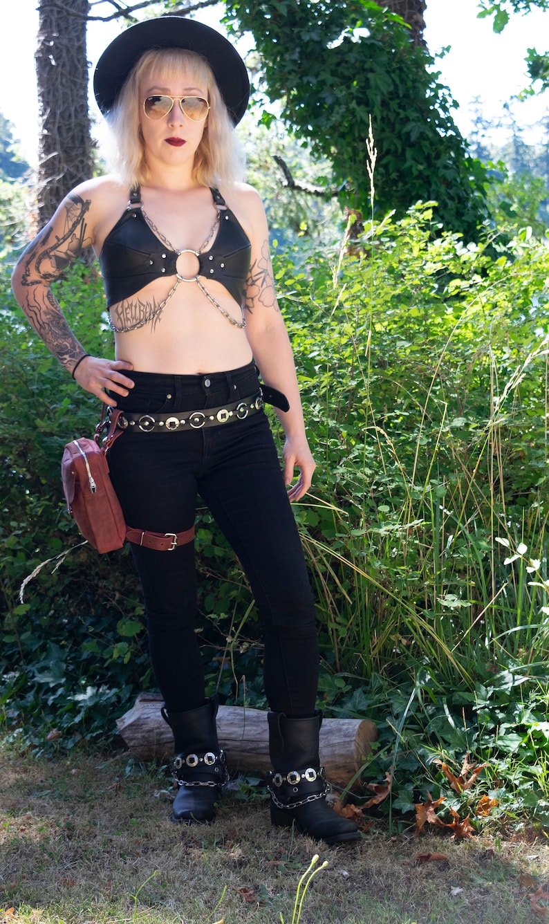 May include: A woman wearing a black leather halter top with a silver chain and ring detail. She is also wearing black jeans, a black belt with silver accents, a brown leather fanny pack, and black boots with silver chain and buckle accents.