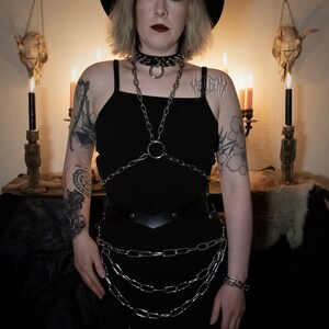 May include: A person wearing a black dress with a chainmail top and a black hat with red candles on top. The person has a tattoo on their arm and is standing in front of a dark background.