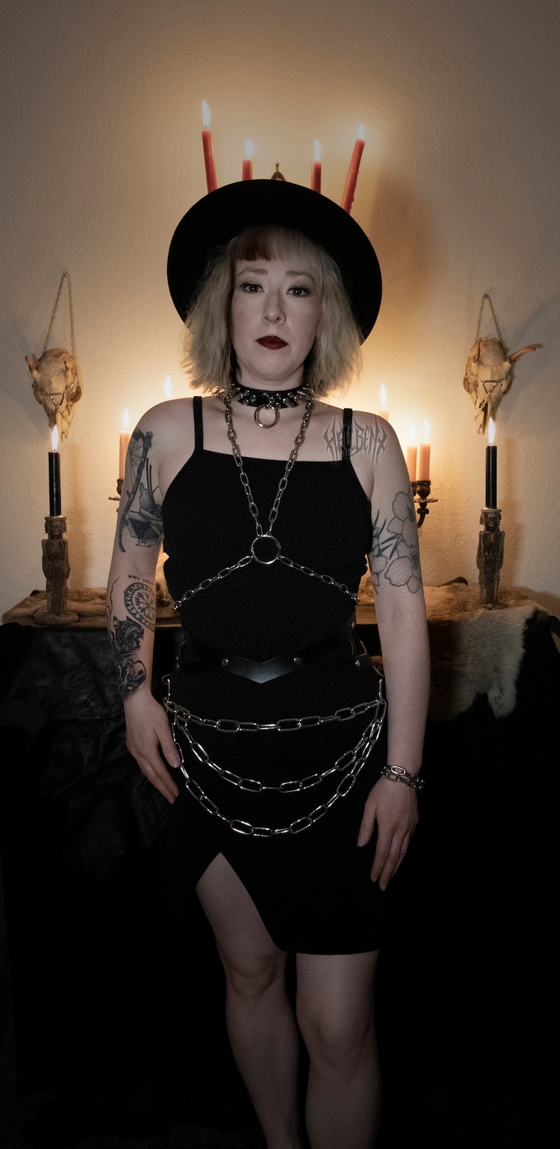 May include: A person wearing a black dress with a chainmail top and a black hat with red candles on top. The person has a tattoo on their arm and is standing in front of a dark background.