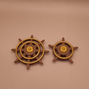 Op de afbeelding: Twee bruine scheepsstuurwielen met goudkleurige accenten op de buitenste ringen en een centraal rond embleem met een gouden letter Q. Het grotere wiel heeft acht spaken, terwijl het kleinere wiel er vijf heeft, beide met een matte afwerking.