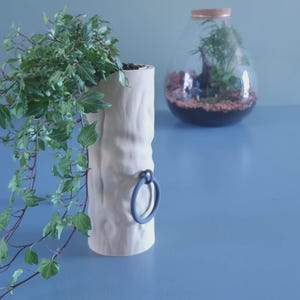 May include: A cylindrical off-white planter with a textured surface resembling tree bark, featuring a single dark gray circular ring handle attached to the side. A trailing green ivy plant grows from the top opening of the planter.