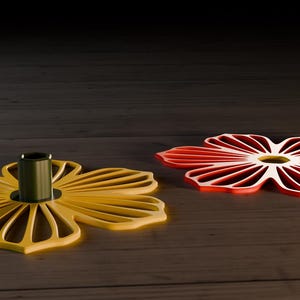 Op de afbeelding: Twee bloemvormige onderzetters. Eén is geel met een donkergroene middenpaal en de andere is rood. De onderzetters zijn gemaakt van een stevig materiaal en hebben een sleufontwerp. Ze zijn op een houten oppervlak geplaatst.