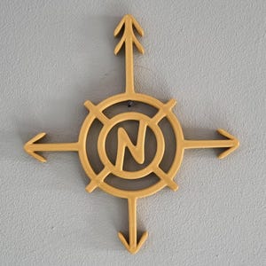 Op de afbeelding: Een goudkleurige kompasroos met de letter "N" in het midden, die het noorden aangeeft. Het kompas heeft vier richtingspijlen en is bevestigd aan een lichtgrijze muur. Het ontwerp is eenvoudig en modern.