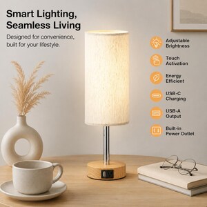 May include: A cylindrical table lamp with a cream-coloured fabric shade, a chrome stem, and a light wood base featuring a built-in power outlet and USB port. The lamp is illuminated, casting a warm glow, and is surrounded by a ceramic mug and glasses.