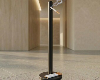 May include: A black metal ashtray with a tall, cylindrical design. A lit cigarette emits a stream of smoke. Two unlit cigarettes rest on the circular base. The ashtray is designed for outdoor or public use.