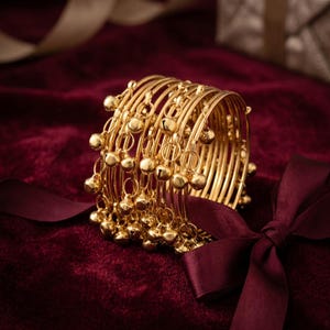 May include: A stack of gold bangles with small dangling bells. The bangles are arranged on a deep red velvet surface, with a burgundy ribbon tied in a bow. The bangles have a shiny, reflective surface.
