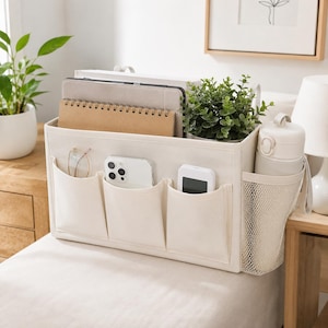 May include: A cream-colored fabric bedside storage caddy featuring three front pockets, a side mesh pocket, and a large main compartment. It holds a tablet, a spiral notebook, a smartphone, a small electronic device, and a water bottle.