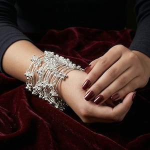 May include: A collection of silver bracelets with small, round beads and floral-like accents. The bracelets are stacked on a wrist, with a dark background and a burgundy velvet surface.
