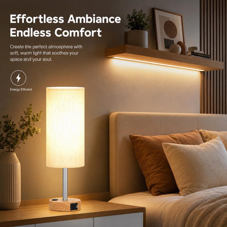 May include: A cylindrical table lamp with a light-coloured fabric shade, a silver metal stem, and a round wooden base featuring a black power switch and a USB port. The lamp emits a warm glow, illuminating the surrounding area in a bedroom setting.