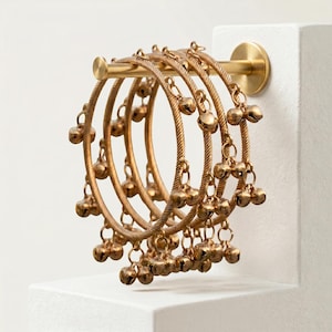 May include: A set of gold-colored bangles with small bell charms hanging from each. The bangles are displayed on a gold bar attached to a white surface. The bangles have a textured, twisted design.
