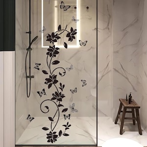 May include: A glass shower door features a black floral vine design with flowers and butterflies. The shower door is set against a marble-patterned wall. A small wooden stool with toiletries sits in the corner.