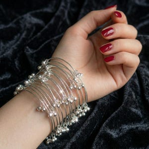 May include: A set of silver bangles with small, dangling bells. The bangles are worn on a wrist with red nail polish. The jewelry is displayed against a dark, textured background.
