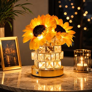 May include: A decorative lamp featuring a cluster of bright yellow sunflowers atop a base constructed from clear, square, faceted glass blocks. The lamp sits on a circular gold-toned metal stand and emits a warm, internal glow from within.