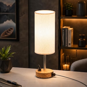 May include: A cylindrical table lamp with a beige fabric shade and a light-colored wooden base. The lamp features a silver metal stem and includes two black USB ports integrated into the base, with a black power cord extending from the side.