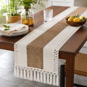 May include: A rectangular table runner featuring a central brown woven panel flanked by two cream-colored textured sections. The runner is finished with a decorative open-weave border and long cream-colored tassels along both short ends.
