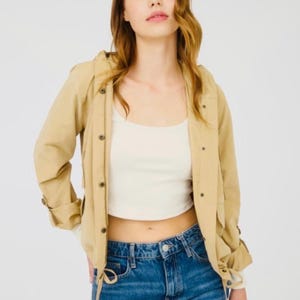May include: A woman wearing a tan hooded jacket over a white crop top and blue jeans. The jacket has button closures and adjustable sleeves. The jeans are a classic blue wash.