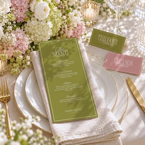 May include: A tall olive green menu card with white text sits on a folded beige linen napkin atop a white plate. Two rectangular place cards, one olive green labeled Theodore Kennedy and one dusty pink labeled Eleanor Smith, are nearby.