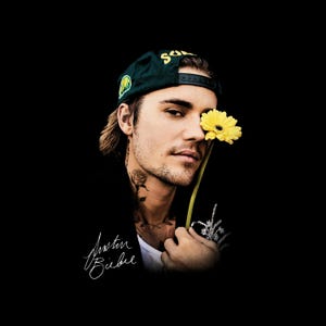 Puede incluir: Una gorra de béisbol verde oscuro con letras amarillas usada por una persona que sostiene una flor amarilla brillante cerca de su rostro. La imagen presenta una firma blanca Justin Bieber sobre un fondo negro sólido, resaltando el detalle floral.