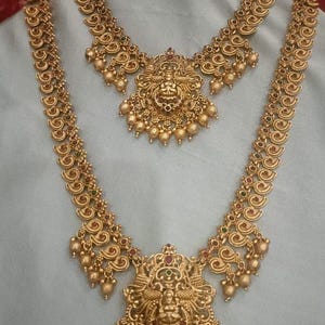 Könnte beinhalten: Ein goldfarbenes Schmuckset bestehend aus einer langen Halskette, einer kürzeren Halskette und einem Paar passender Jhumka-Ohrringe. Jedes Stück zeichnet sich durch filigrane Metallarbeiten, kleine hängende Perlen und zentrale Anhänger aus.