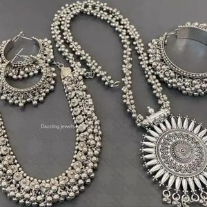 May include: A silver-toned jewelry set featuring a long necklace with a large circular sunburst pendant, a shorter beaded necklace, a pair of hoop earrings with dangling beads, and a wide cuff bracelet. The text Dazzling jewels india is visible.