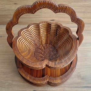 May include: A wooden, collapsible fruit basket with a carved floral design. The basket is made of light brown wood and features a scalloped edge and a handle. The interior has a layered, wave-like pattern. The base is also scalloped.