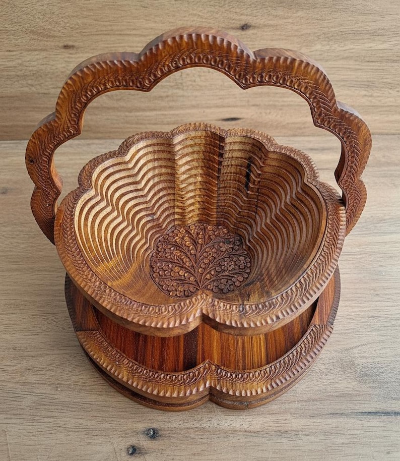 May include: A wooden, collapsible fruit basket with a carved floral design. The basket is made of light brown wood and features a scalloped edge and a handle. The interior has a layered, wave-like pattern. The base is also scalloped.