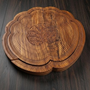 May include: A wooden, collapsible fruit basket with a floral design carved into the center. The basket is made of thin, curved wooden slats and has a scalloped edge. The wood has a warm, brown tone, and the basket is set against a dark wood surface.