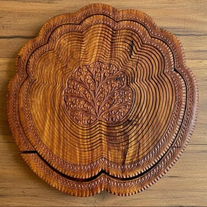 May include: A round, wooden fruit basket with a carved floral design in the center. The basket has a scalloped edge and is made of thin, curved wooden slats. The wood has a warm, brown tone.