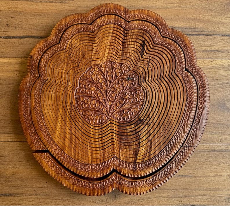 May include: A round, wooden fruit basket with a carved floral design in the center. The basket has a scalloped edge and is made of thin, curved wooden slats. The wood has a warm, brown tone.