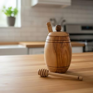 May include: Wooden honey pot with a lid and a honey dipper. The pot is barrel-shaped and made of light brown wood. The honey dipper is lying next to the pot on a wooden surface. Kitchenware.