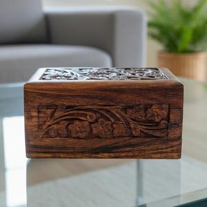 May include: A rectangular wooden box with intricate floral carvings. The box is a rich brown color and sits on a glass surface. The carvings are detailed and cover the top and sides of the box, showcasing a natural wood grain.