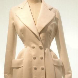 May include: A cream-colored double-breasted coat with a tailored fit. The coat features a notched collar, two front pockets, and six buttons. The coat is displayed on a mannequin.