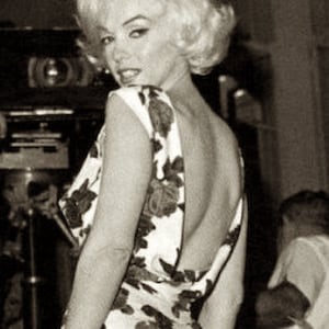 May include: A vintage black and white photograph of a woman in a floral print dress and white heels. She is holding a white clutch and looking over her shoulder. The dress has a low back.