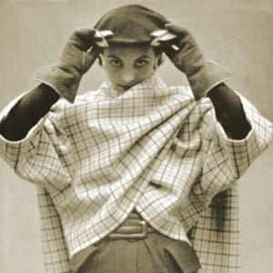 Può includere: Immagine in seppia di una persona con un mantello a quadri, guanti e un berretto. Il mantello è drappeggiato sulle spalle e fissato in vita con una cintura. I guanti hanno polsini svasati. Il berretto è scuro.