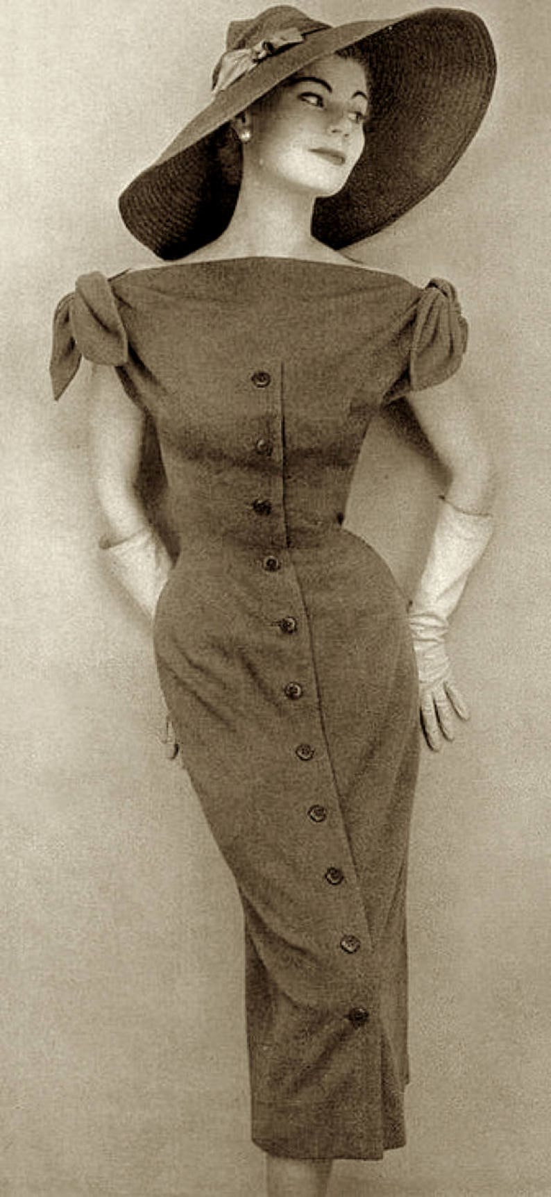 May include: A vintage-style dress with a button-down front and a fitted silhouette. The dress is a muted brown color and features short sleeves with a bow detail. A wide-brimmed hat and white gloves complete the ensemble.