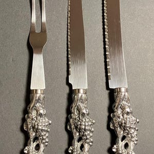 Puede incluir: Un juego de cuchillos para queso plateados con un tenedor, un cuchillo y un cuchillo dentado. Los mangos están intrincadamente diseñados con un motivo de vid. Las hojas son brillantes y el juego se exhibe sobre una superficie oscura.