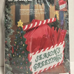 May include: A Bucilla Season's Greetings buffet caddy kit. The kit includes plastic canvas, acrylic yarn, and embroidery floss. The caddy is red, white, and green, with a Christmas tree design and the words "Season's Greetings". The caddy measures 9.5" x 10" x 7".