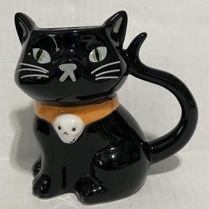 May include: A glossy black ceramic mug shaped like a sitting cat. The cat has green eyes, white whiskers, and a nose. It wears an orange collar featuring a white skull charm. The cat's tail forms the handle of the mug, curving upward and outward.