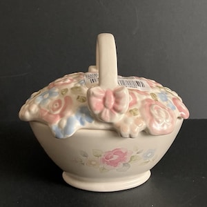 Può includere: Un contenitore in ceramica a forma di cestino con coperchio, manico e fiocco rosa. Il coperchio è decorato con fiori rosa, blu e bianchi scolpiti. La base del contenitore presenta disegni floreali dipinti. La combinazione di colori è pastello.