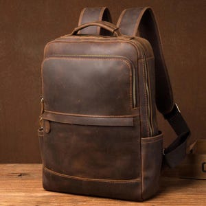 May include: A brown leather backpack with multiple zippered compartments and adjustable shoulder straps. The bag has a rectangular shape with a front pocket and a top handle. The leather has a distressed look, giving it a vintage appearance.