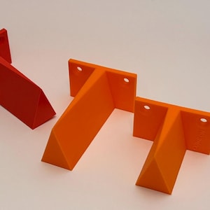 May include: Three geometric, T-shaped brackets in shades of red and orange. Each bracket has a rectangular top with two holes and a triangular support. The brackets are arranged on a white surface.