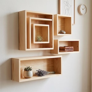 Könnte beinhalten: Eine Sammlung hellfarbiger, schwebender Holzregale in verschiedenen quadratischen und rechteckigen Formen. Einige Regale enthalten kleine Pflanzen, einen Stein und Bücher. Die Regale sind an einer weißen Wand befestigt und schaffen eine moderne, minimalistische Ästhetik.