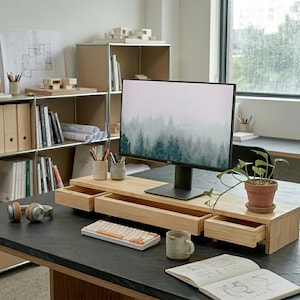 May include: A light-colored wooden monitor stand with three drawers, holding a computer monitor displaying a forest scene. A small potted plant, keyboard, and mug are on the desk. Shelves with books and office supplies are in the background.