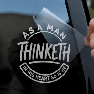 May include: A white vinyl decal featuring the text AS A MAN THINKETH IN HIS HEART SO IS HE arranged in a circular design. The decal is partially peeled away from its clear transfer tape, revealing the adhesive side against a dark surface.