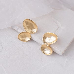 Peut inclure: Une paire de boucles d'oreilles dorées à la forme organique unique. Chaque boucle d'oreille présente trois pièces rondes texturées qui se chevauchent.