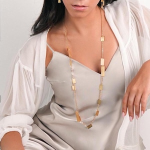 Könnte beinhalten: Eine Frau trägt eine weiße Bluse und ein beiges Schlupfkleid mit einer goldenen Kettenkette und passenden Ohrringen und Ring. Sie sitzt auf einem Holzstuhl mit gesenktem Kopf.