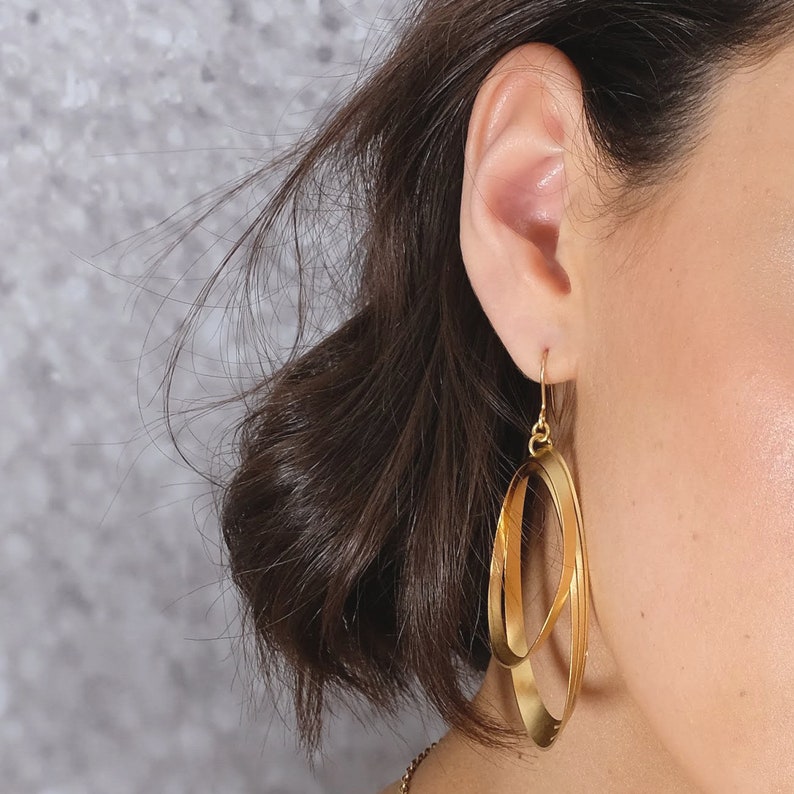 May include: Gold hoop earrings with a double-loop design. The earrings are dangling from the earlobe.